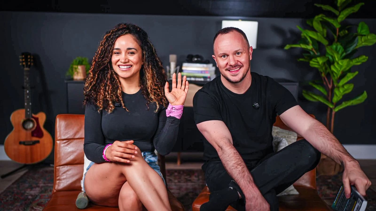Veronica waving and Stephen smiling, seated together in their studio